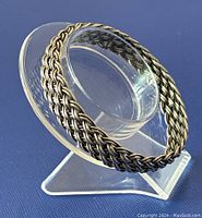 Braided silver bangle displayed on clear stand showing the textured woven design and thickness.
