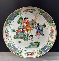 Front view of the round shallow porcelain plate showing hand painted traditional scene in center and famille verte floral border with gold rim.