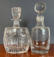 Two crystal decanters side by side, one clear crystal with rounded stopper and a label, the other a heavier cut crystal with a faceted stopper