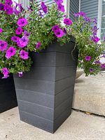 Front and side views of a single black resin planter filled with purple petunias and green foliage placed on a concrete area outside a house.
