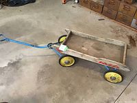 Photo of entire wooden wagon showing rectangular wooden bed, blue metal frame, handle, and yellow wheels on a concrete floor.