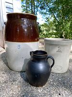 Number 6 two-tone brown and beige pickling crock, number 2 Imperial Medalta crock, and black crockery pitcher grouped together on concrete ground.