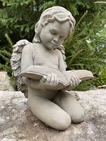 Front right angled view of resin angel statue kneeling with open book, outdoors on rock with greenery background.