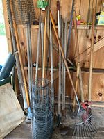 Overall view of garden tools leaning against wall including post hole digger, cultivators, rakes, shovels, and roll of wire fencing