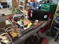Overall view of table with wire shelf holding boxes of assorted household tools, knobs, mailbox, and hardware