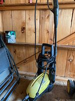 Side view of yellow Karcher pressure washer showing handle, wheels, and hose wrapped around it, placed indoors against wooden wall.
