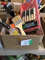 Box filled with multiple paintbrushes of various sizes and bristle types, a foam brush set in red packaging, and a paint roller pan.