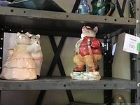 Shelf display of two ceramic novelty teapots: cats dancing and pig with walking stick by Fitz and Floyd.