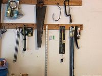 Wide view of wooden wall-mounted rails with a vintage hand saw, a hacksaw, measuring stick, magnifying glass, and hooks.
