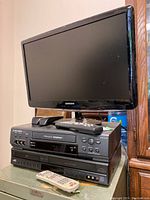 Samsung HDTV monitor, Mitsubishi VHS player, JVC DVD player, and remotes stacked on top of each other on small table.
