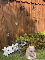Photo of a black metal wire garden trellis, white ornate metal garden edging panels, metal garden wall flowers mounted on wooden fence, and heavy concrete pig statue on grass.
