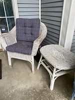 White painted wicker chair with grey cushions and matching table on porch floor against siding wall.