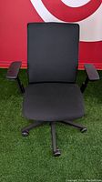 Front view of the Steelcase Think V2 office chair showing black fabric seat and backrest with adjustable armrests and caster wheels base.