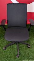 Front view of one black cushioned Steelcase Think V2 office chair with armrests and caster five-wheel base on green carpet
