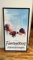 Framed "Firecrackers" movie poster in black frame leaning against wall on wooden floor.