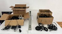 Two cardboard boxes filled with tangled black and some white electrical cables and charger cords, placed on white table