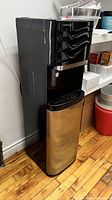 Full view of the water cooler showing black glossy dispensing section and stainless steel bottom section on wood floor.