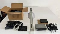 Overview photo showing multiple black Ethernet switches, Cisco router, white vertical Ethernet switch, their power adapters, and cables on a white table