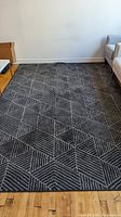 Full view of dark gray and light gray geometric patterned rectangular rug placed on wooden floor next to sofa and white wall.