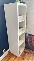 Side view of white shelving unit standing against a blue wall and white brick wall with hardwood floor and electrical outlets visible.