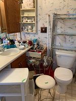 View of bathroom corner with wooden vanity cabinet, plastic drawer unit, toilet, over-toilet metal storage with glass shelves, metal vanity seat, and various bathroom accessories on sink and wall cupboard.
