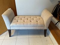 Bench full view showing light beige tufted upholstery and dark wooden legs in tile floor setting