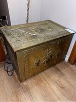Full view of the rectangular brass chest with embossed ship patterns on the front and lid, sitting on the floor.