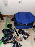 Lot contents including Nikon D50 DSLR with lens, FujiFilm Finepix S camera with Sigma lens, Nippon Kogaku Nikkor lenses, Nikon SB 400 flash, Conquest tripod, cords, universal converter, and blue camera bag placed on carpeted floor.