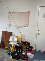 Photo showing various camping equipment including coolers, fly fishing rod, flashlight, and other accessories arranged by a wall and door.