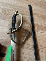 Photo showing full length of ceremonial sabre alongside sheath, displaying blade, brass guard, black handle with wear, and braided tassel.