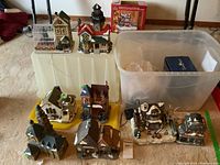 Eight Christmas village buildings arranged with two plastic storage containers beside them.
