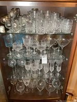 Wide front view of multiple shelves inside china cabinet showing an assortment of clear glass and crystal stemware and bar glasses.