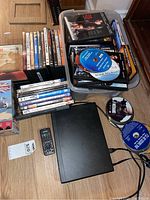 Photo of Philips Blu-ray/DVD player with remote, various DVDs stacked and loose discs visible, and box set VHS tapes on wooden floor.