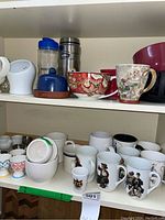 Shelf with various kitchen bowls, mugs including Norman Rockwell themed mugs, thermos, butter bowl with lid, and decorative kitchen items.