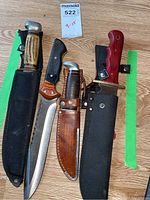 All four hunting knives displayed with sheaths on a wooden floor, showing overall size and variety.