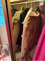 Photo showing jackets on hangers inside a closet, including a tan suede jacket with faux fur lining, black belted jacket, cream fleece jacket, and pink jacket.