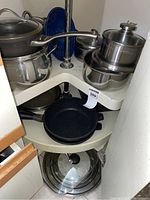 Two shelves in a corner cupboard filled with assorted pots, pans, lids, and a blue colander. Visible are mostly stainless steel cookware and a black frying pan.