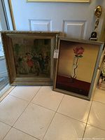 Photo of two framed artworks leaning against a door: a vintage style oil painting and a rose print.