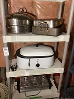 3-shelf plastic shelving unit with electric roasting pan on middle shelf, stainless steel pans above, and warming station below