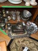 Overview photo showing a variety of silver plated and aluminum serving dishes, trays, and bowls stacked on a wooden shelf and floor with visible tarnish.