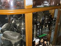 View of glass shelf display showing silver plated covered butter dish, teapots, and salt and pepper shakers with tarnish