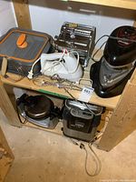 Overview of two wooden shelves holding a variety of untested small kitchen appliances including a hand mixer, pasta maker, waffle maker, deep fryer, and other equipment.
