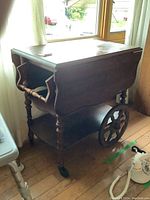 Full view of the tea wagon showing the drop leaf sides, turned legs, large wooden wheels on one side, and caster wheels on the other.