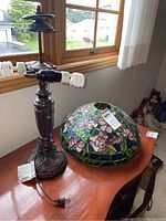 Photo showing the full lamp with metal base and separate Tiffany style floral stained glass dome shade on a wooden surface by window.