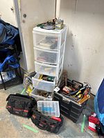 Overall view of five-drawer white plastic storage container with tools inside and two black tool bags with a small plastic container of screws or nails on the floor.