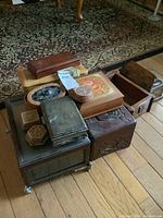 Photo showing assorted wood and metal trinket boxes stacked together on a wooden floor, highlighting various sizes and shapes.
