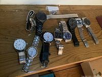 Full view of nine men's wrist watches arranged on a wooden surface showing variety of styles and bands.