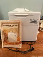 Bread maker machine with the cookbook leaning on it, showing the full unit and the cookbook cover.
