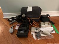 Photo showing the Nikon Coolpix camera, Samsung flip phone with charger, binoculars, silver tripod, and carrying cases arranged on floor.