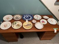 Full view of 12 assorted plates laid on desk showing varied designs including scenic, floral, bird motifs and different shapes.